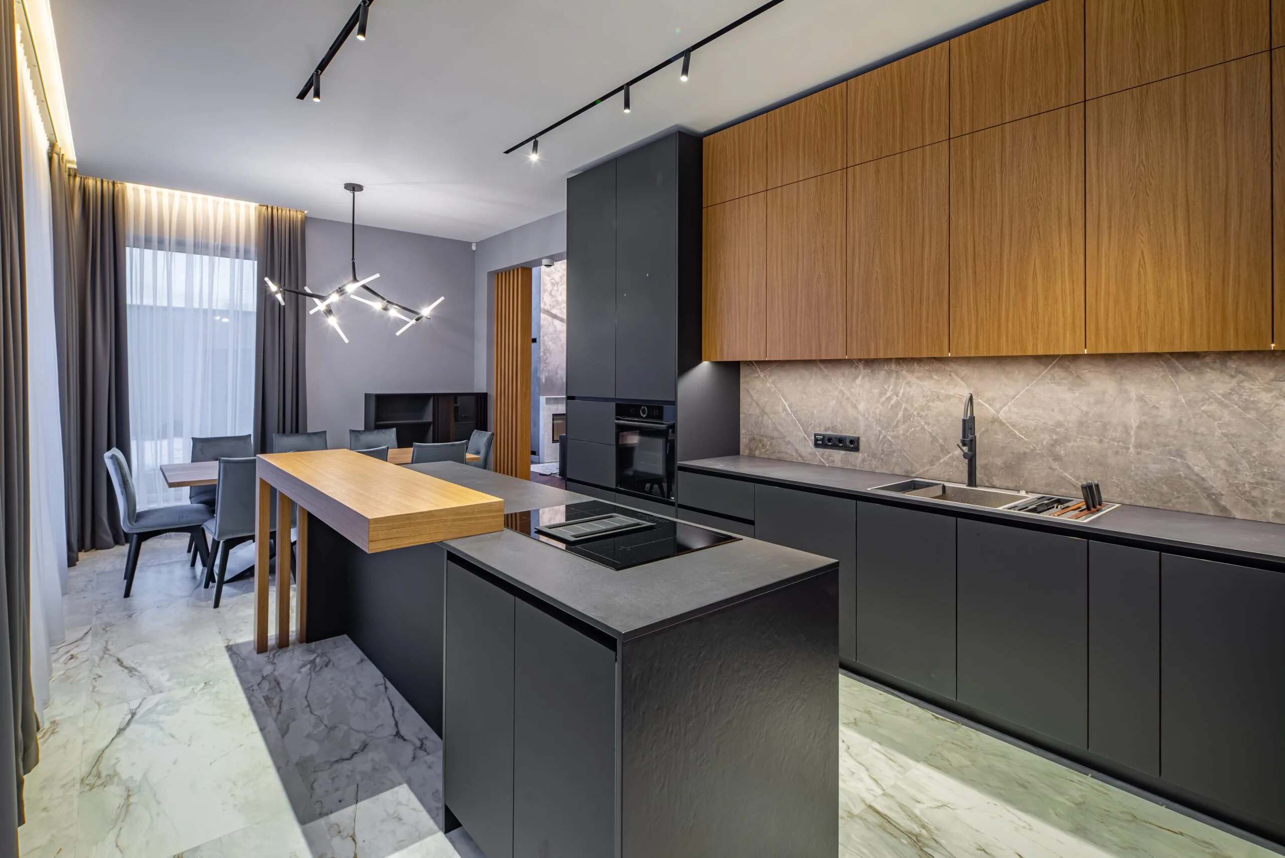 shutterstock_1932352316-scaled-1-jpg Modern kitchen with black cabinets, marble countertops, a wooden island, and an industrial-style light fixture. Dining area with chairs and a table in the background.